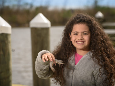 Girl, Smiling, Holding A Whole Shrimp, On A Pier With Water And Land In Background.